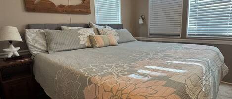 A bedroom with a bed, nightstand, lamp, ceiling fan, and large windows with blinds.