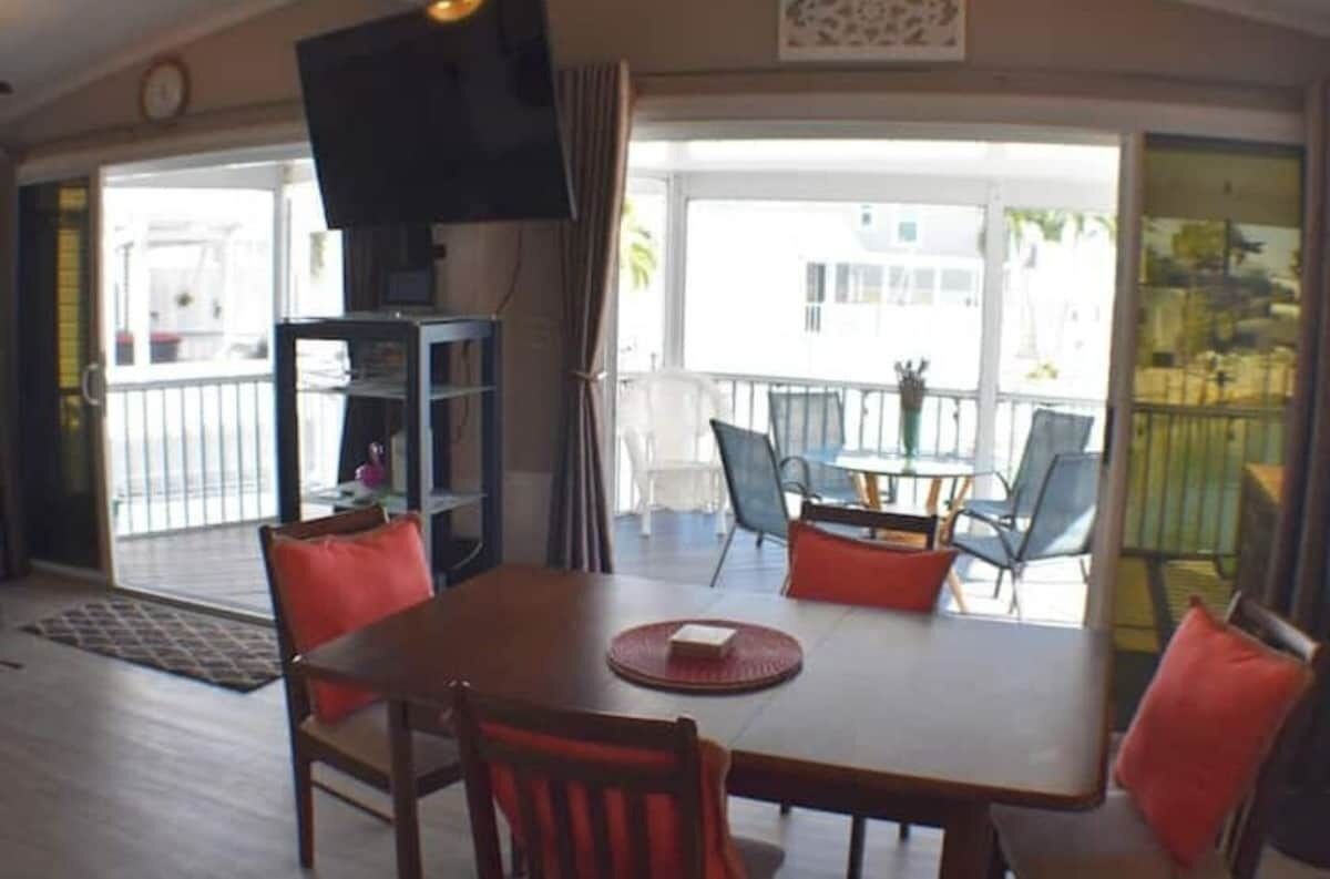 Un coin repas avec une table et des chaises, un téléviseur posé sur un meuble, et une vue sur le balcon extérieur.