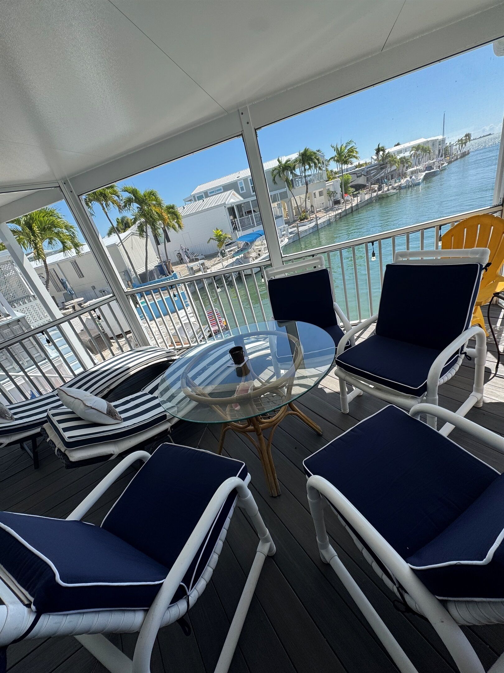 Une terrasse dotée de chaises longues et d’une table en verre, offrant une vue sur une marina avec des bateaux et des palmiers.