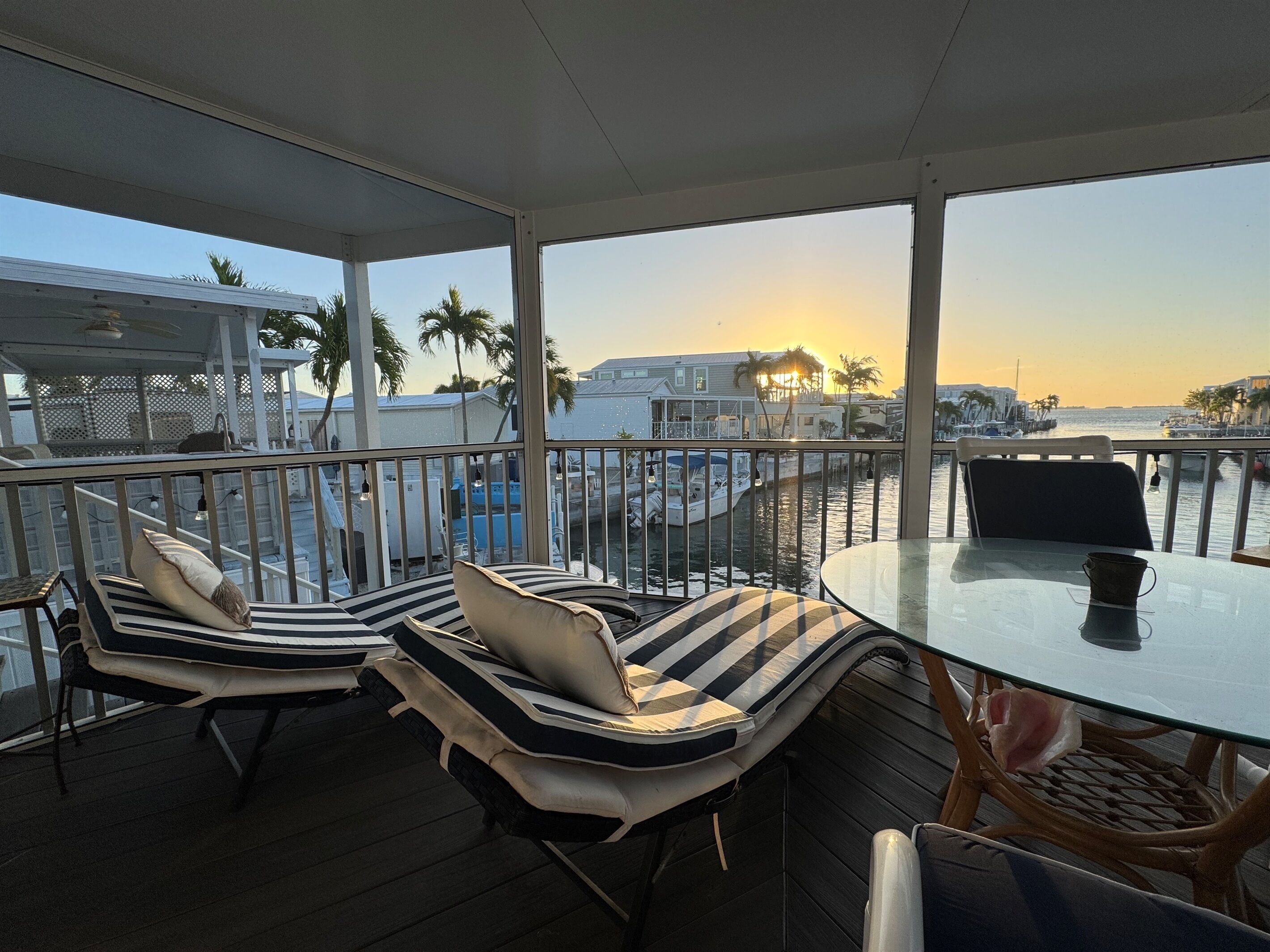 Un espace extérieur am énagé, abrité et doté de fauteuils et d’une table, offrant une vue sur une marina avec des bateaux et des palmiers.
