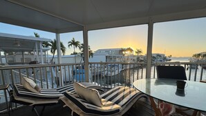 A covered outdoor seating area with lounge chairs and a table, overlooking a marina with boats and palm trees. - Pelican's Roost, Waterfront comfort at Venture Out (Cudjoe Key)