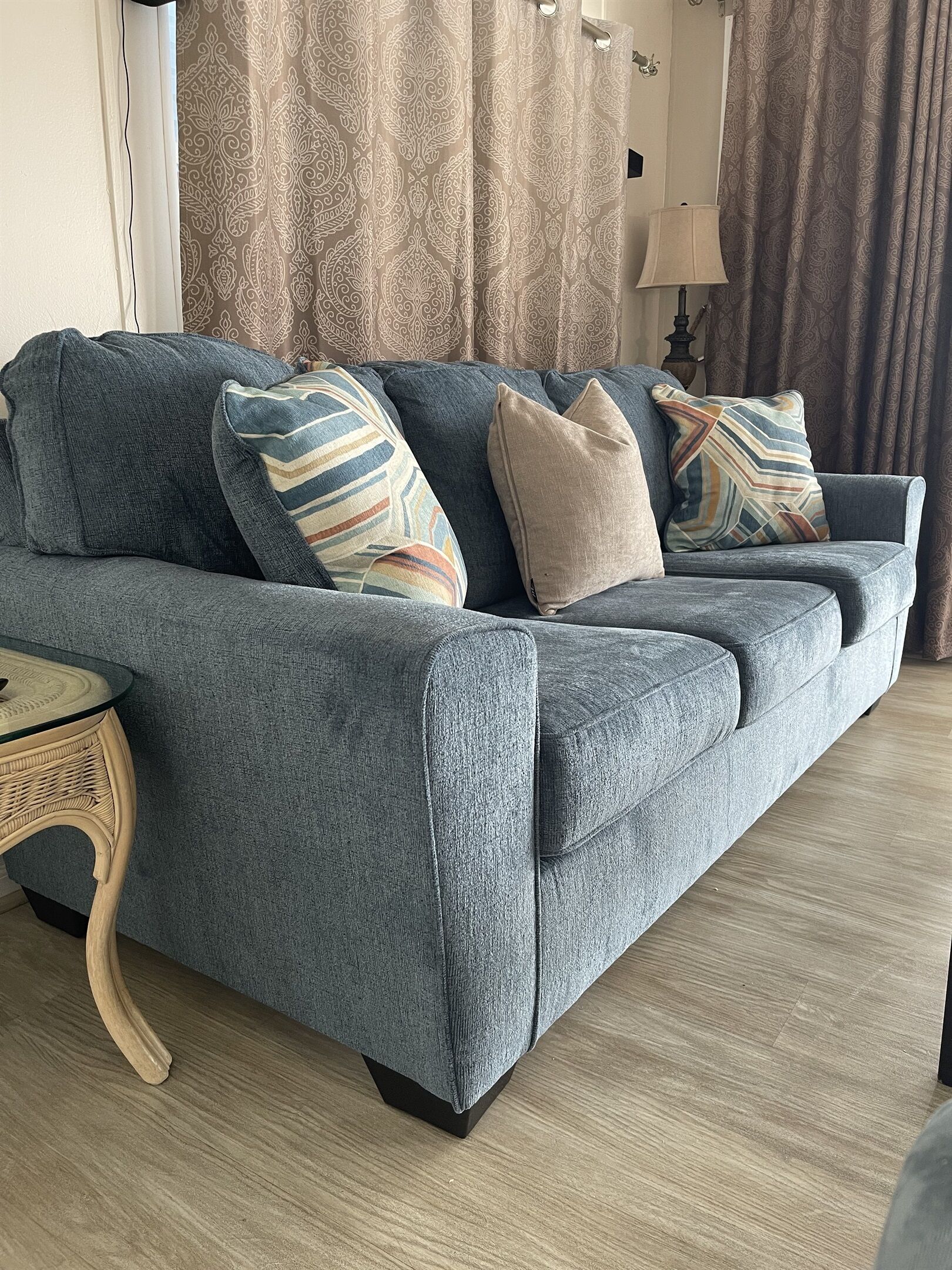 A grey sofa with multiple pillows, a wicker side table, and a lamp.