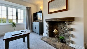 A room with a stone fireplace, a television on a cabinet, a window with curtains, and a table with a remote control. - Curzon House 2 (Cleethorpes)
