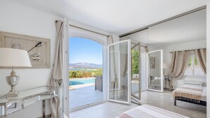 Un dormitorio con una cama grande, un ventanal de vidrio que da a un balcón con piscina y vistas a montañas.