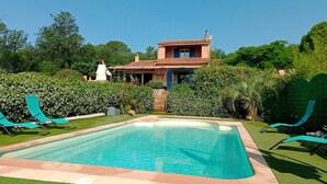 A pool with lounge chairs, a house with a tiled roof, and lush greenery.