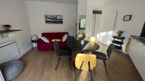 Un espace de vie compact comprenant un canapé rouge, une table à manger avec des chaises, un petit coin cuisine et une étagère fixée au mur.
