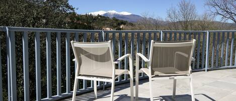 Two white chairs on a balcony with a metal railing, overlooking a snow-capped mountain and trees.