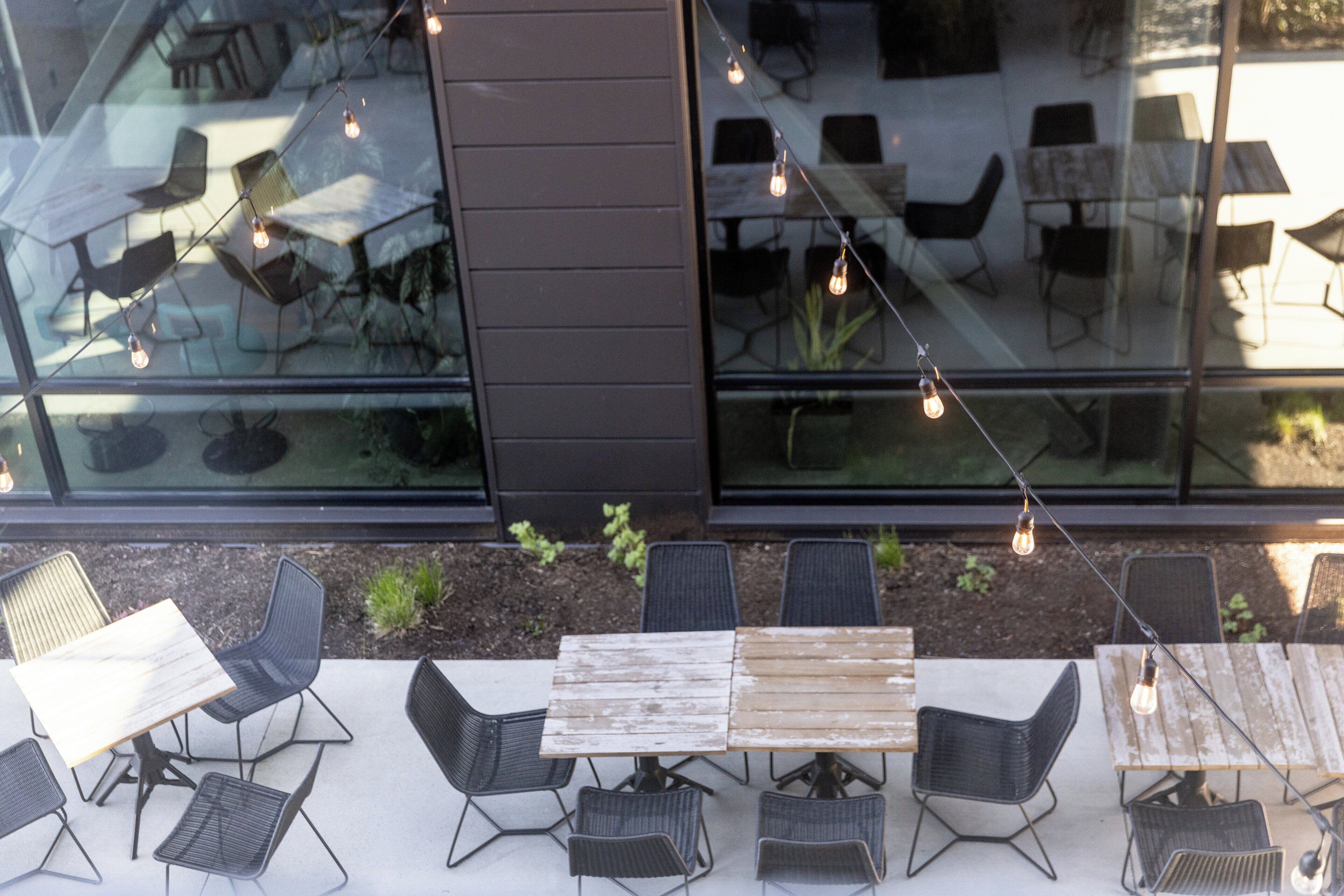 An outdoor seating area with tables and chairs, string lights, and a view of the interior through glass walls.