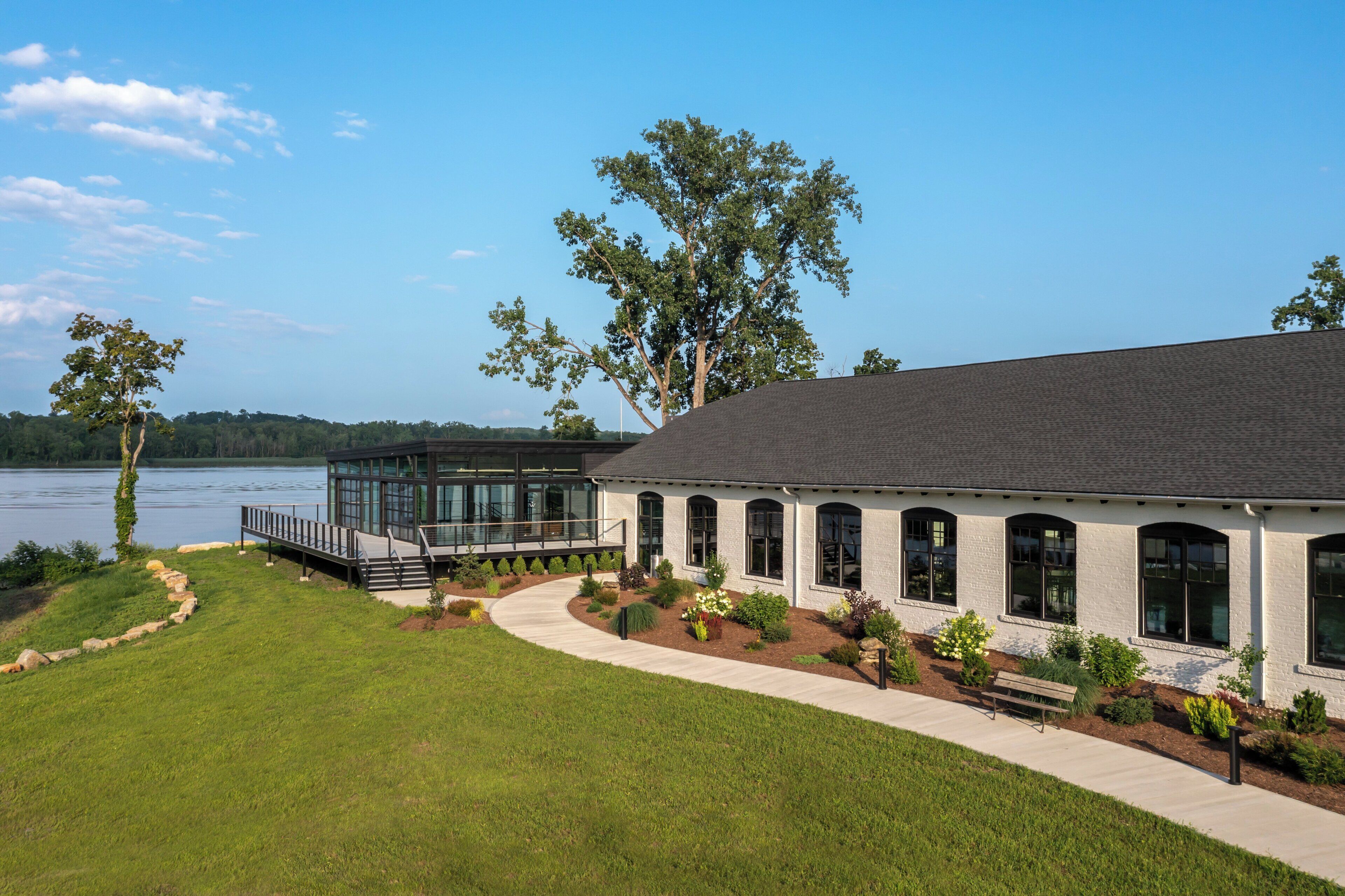 A modern house with a large deck overlooking a lake, surrounded by well-maintained lawns and trees.