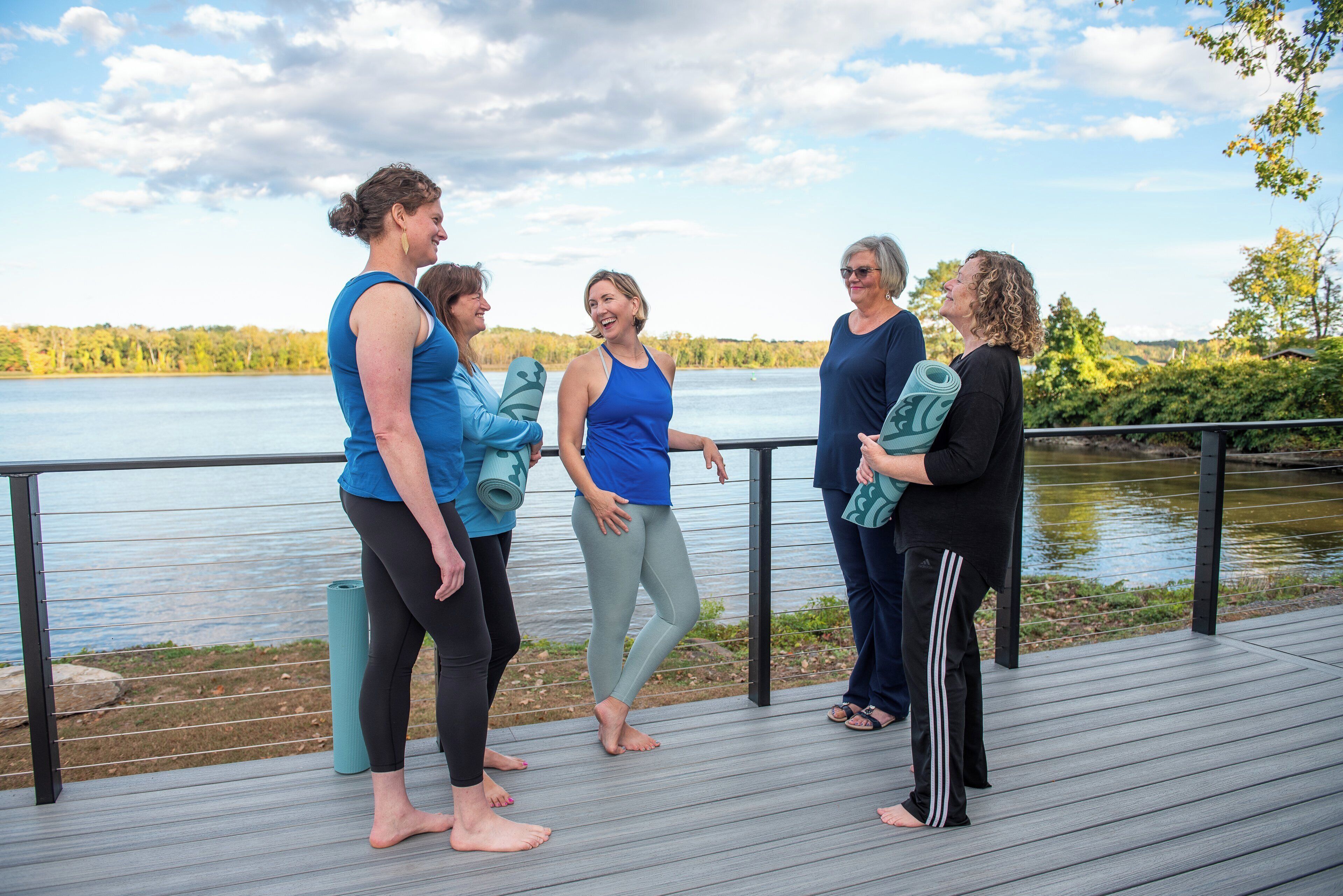 A group of people on a wooden deck by a lake, some holding yoga mats.