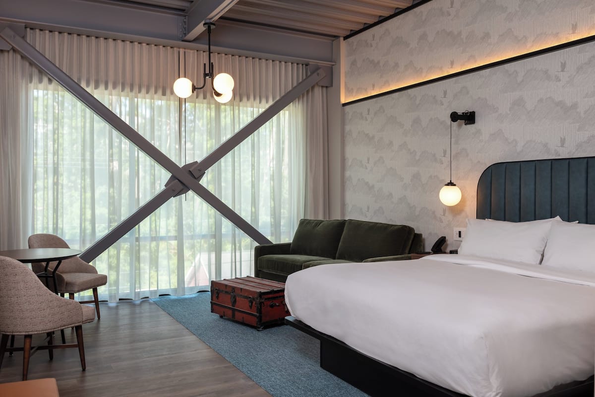 Een hotelkamer met een groot bed, een groene bank, een kleine tafel en een stoel. Er is een raam met gordijnen en uitzicht op bomen.