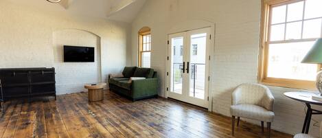 A spacious room with wooden flooring, a green sofa, a black cabinet, and a television mounted on a white brick wall.