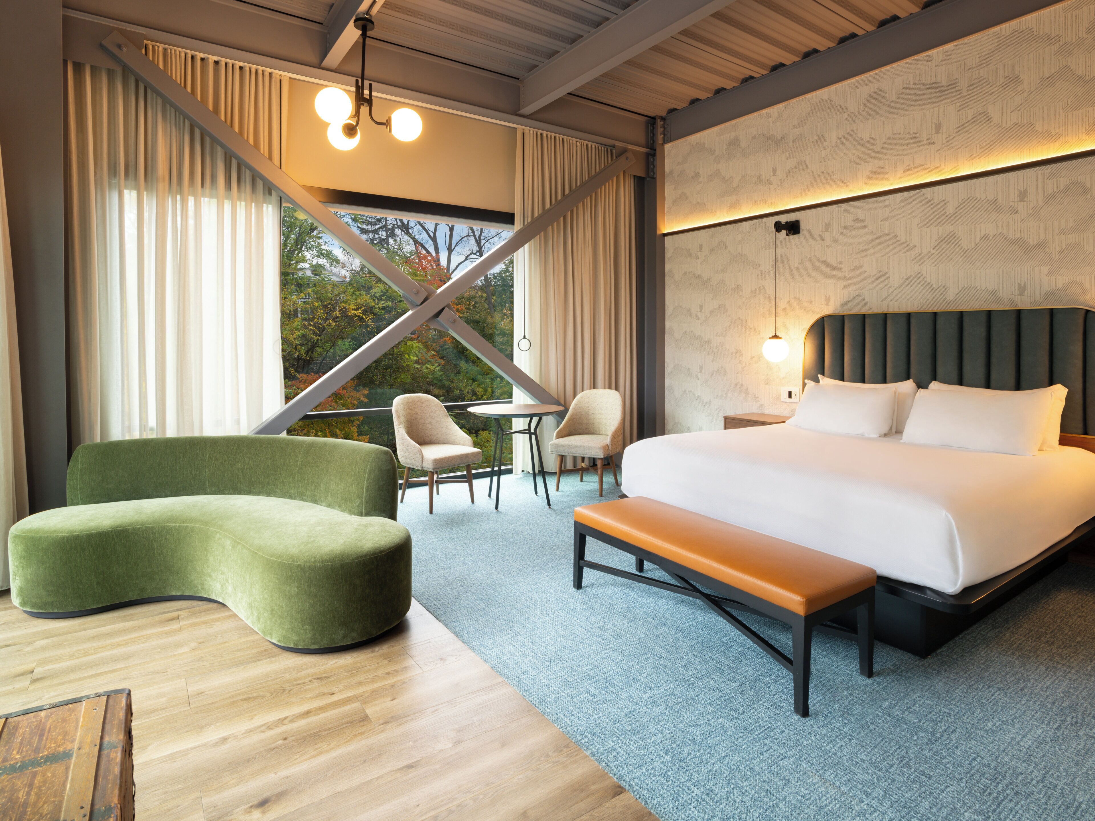A modern hotel room with a large bed, a curved green sofa, two armchairs, a small table, and a view of trees through large windows.