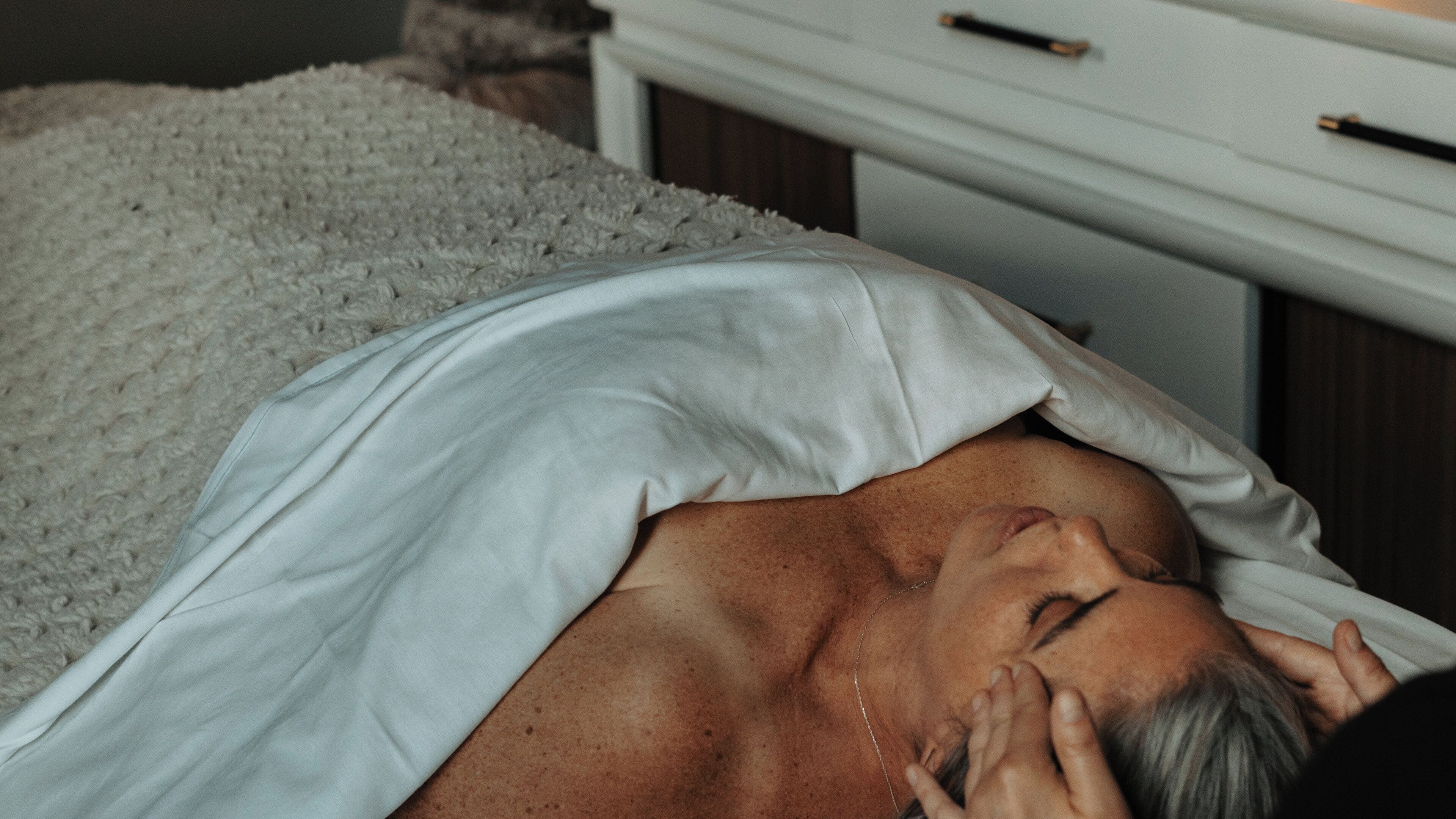 A person lying on a bed with a white blanket, covered by a white pillow.