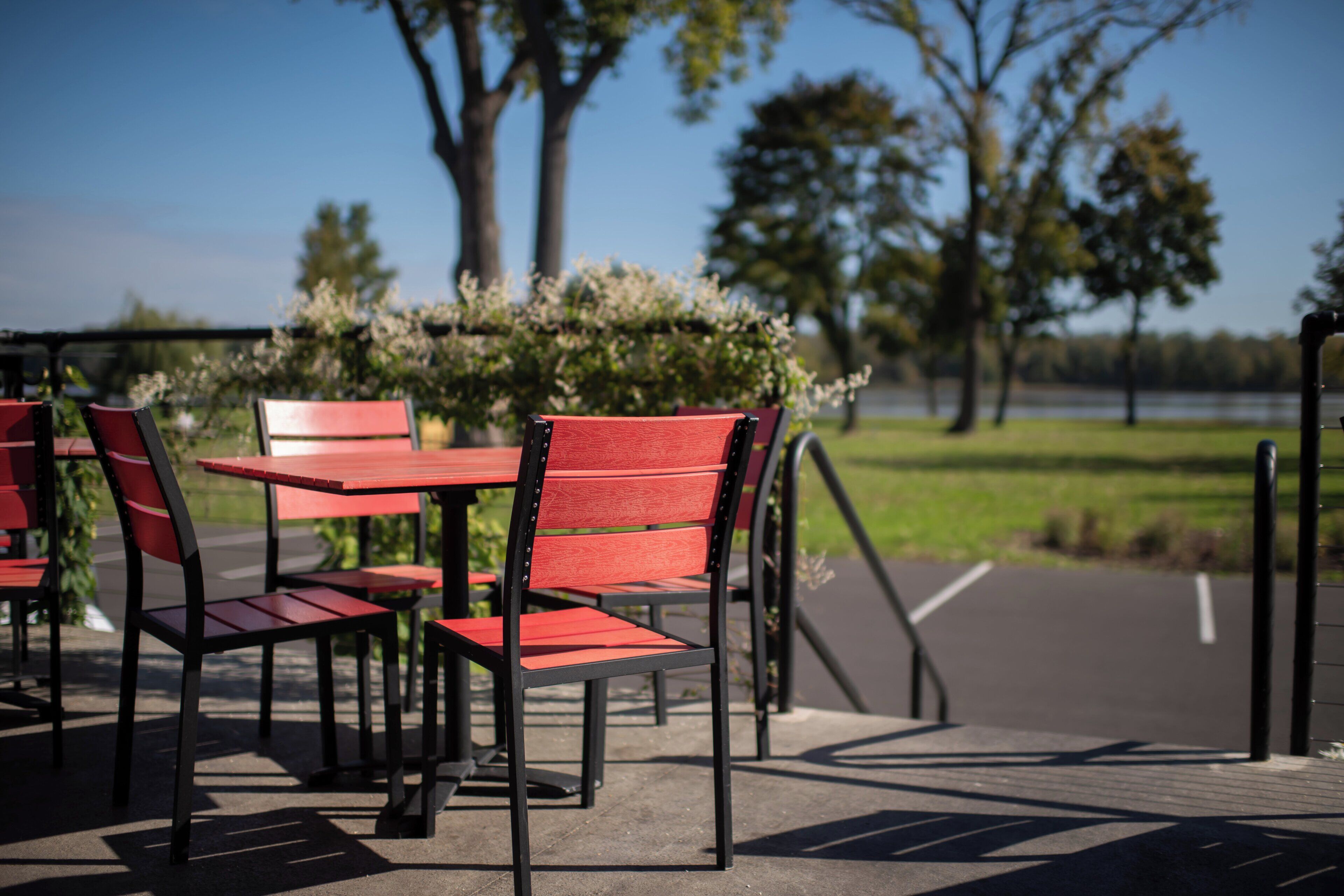 Outdoor seating with tables and chairs, a paved area, and a view of trees and a body of water.