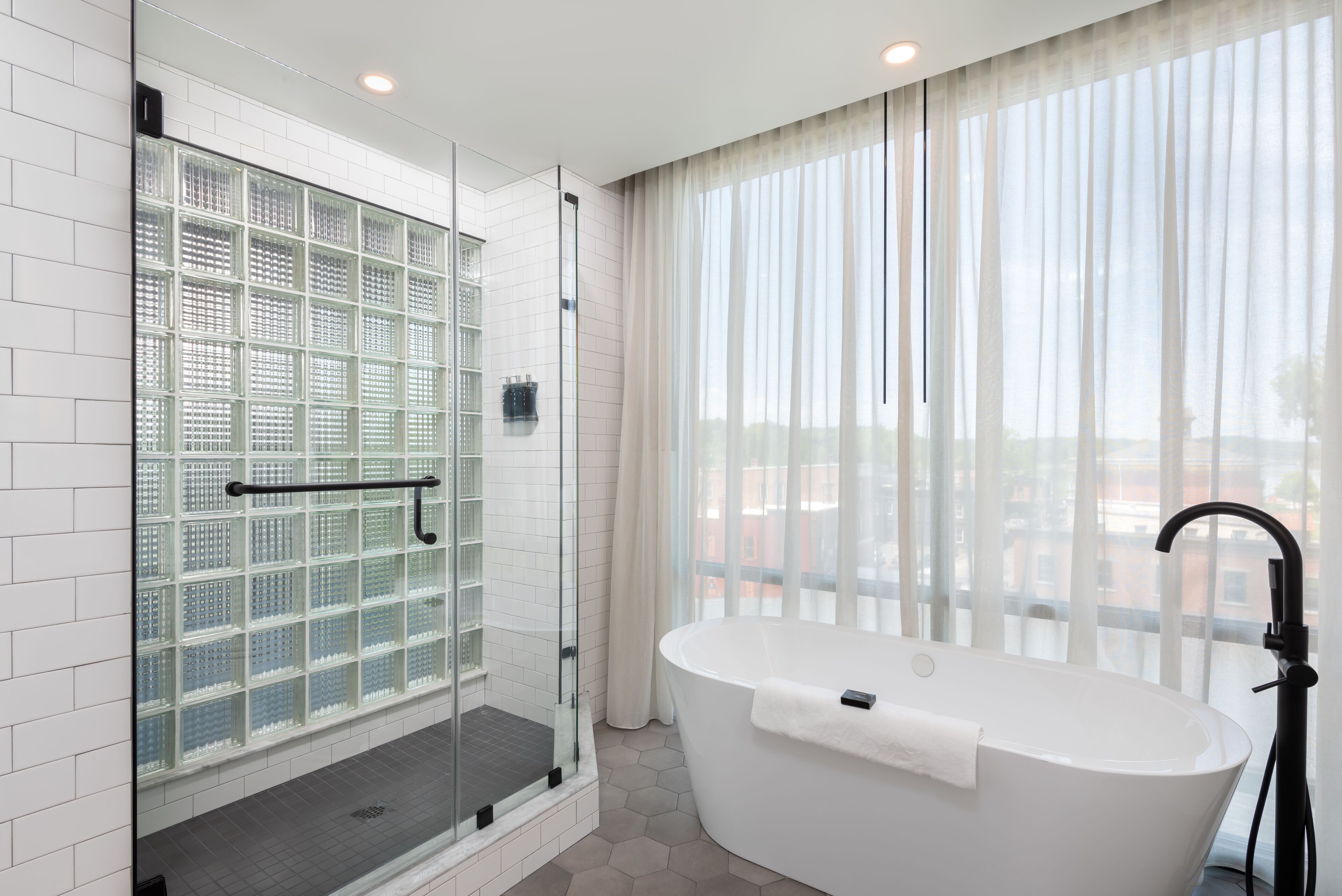 Ein modernes Badezimmer mit einer Glaskabine, einer freistehenden Badewanne und einem großen Fenster mit durchscheinenden Vorhängen.