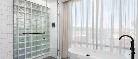 Ein modernes Badezimmer mit einer Glaskabine, einer freistehenden Badewanne und einem großen Fenster mit durchscheinenden Vorhängen.