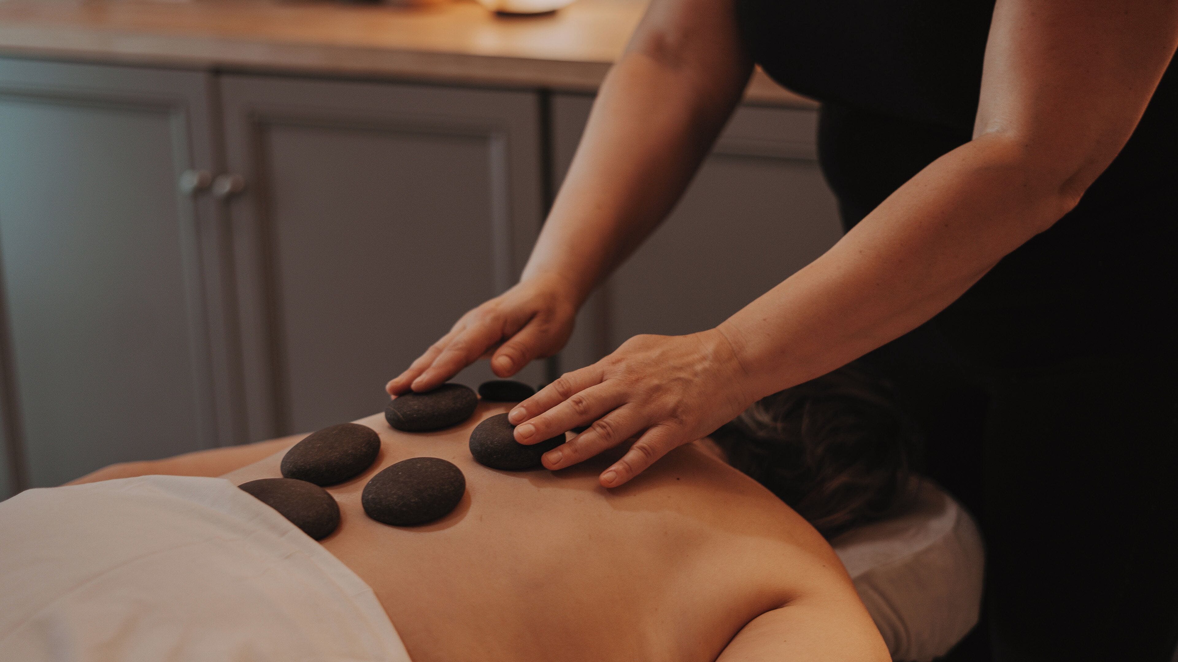 A massage therapy session with a person lying face down receiving pressure from another individual using stones.