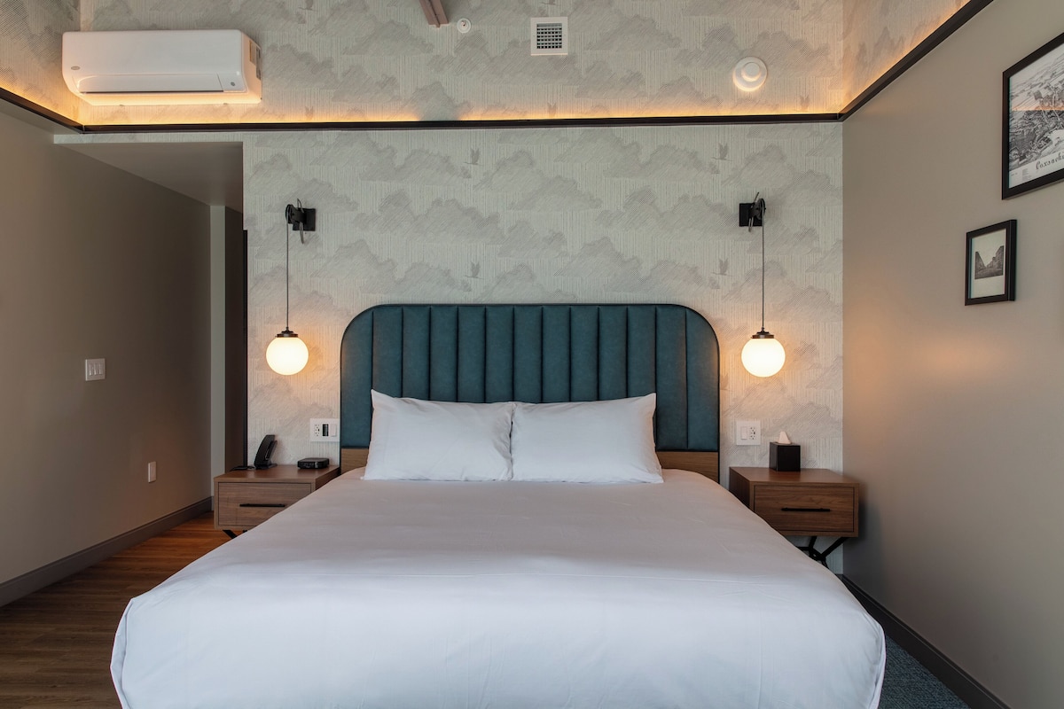 Een net opgemaakt bed met hoofdbord, twee nachtkastjes met lampen, en een wandairconditioner.