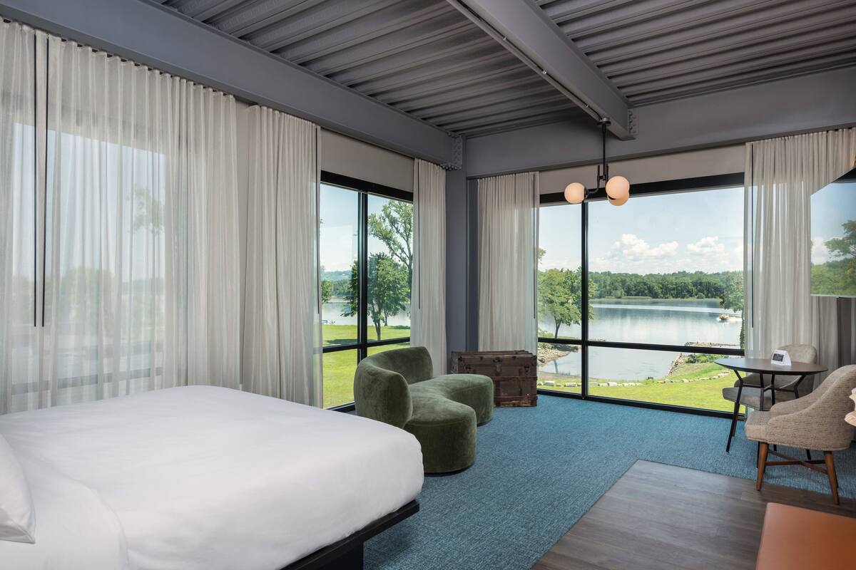 Een moderne hotelkamer met een groot bed, groene fauteuils, een klein tafeltje en uitzicht op een meer.