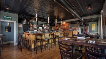 A bar area with wooden chairs and tables, a bar counter with bottles, and a ceiling with exposed pipes.