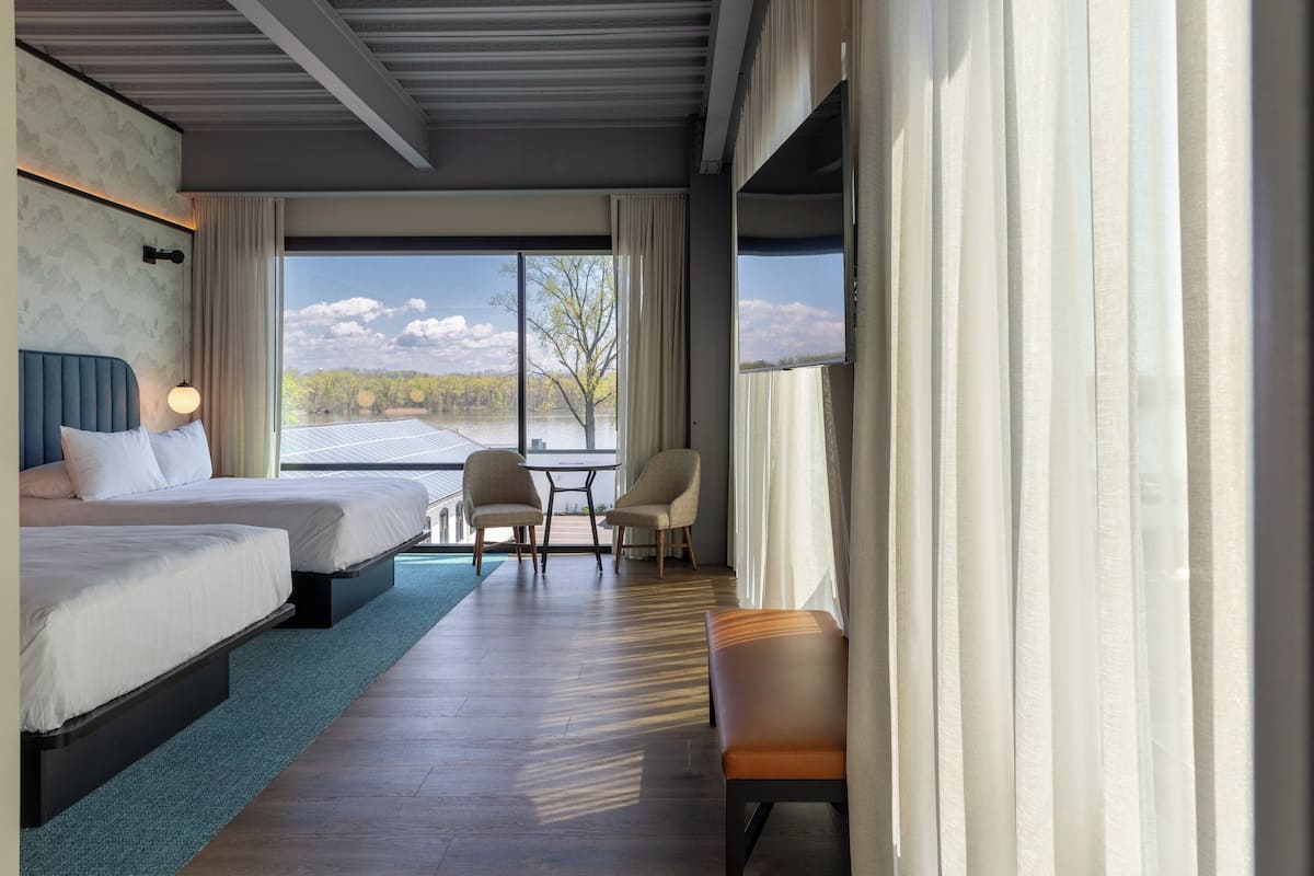 Een hotelkamer met twee bedden, een zithoek met stoelen, uitzicht op een meer en een groot raam met gordijnen.