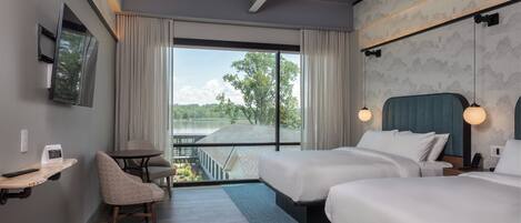 Habitación de hotel con dos camas, un escritorio, una silla, un banco, un televisor y un ventanal con vistas a un lago.