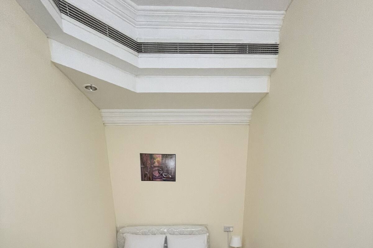 A narrow hotel room with a bed, a nightstand, a lamp, and a small painting on the wall.