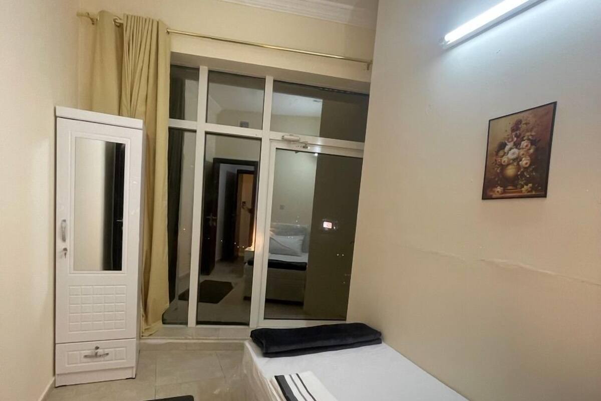 A small, well-lit bedroom with a bed, a black mat, a wardrobe, and a sliding glass door leading to a bathroom.