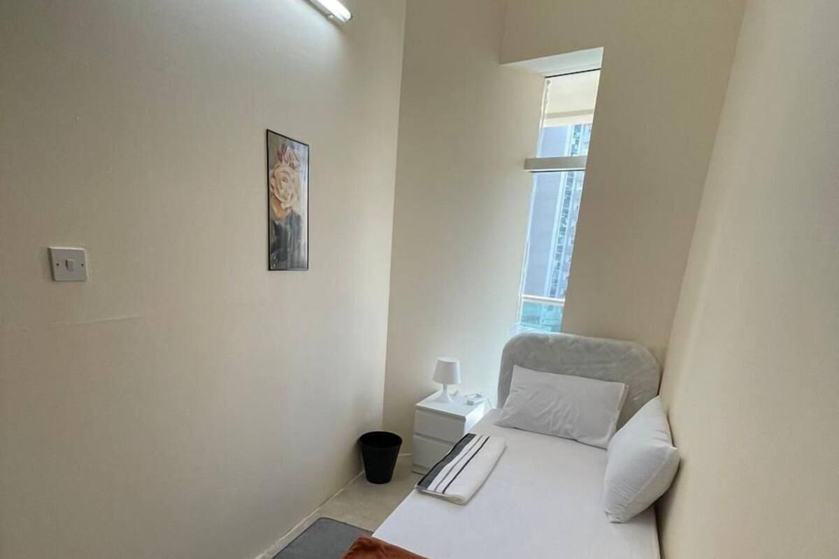 A small, neatly arranged bedroom with a single bed, a bedside table, a nightstand, a lamp, a window, and a wall-mounted painting.