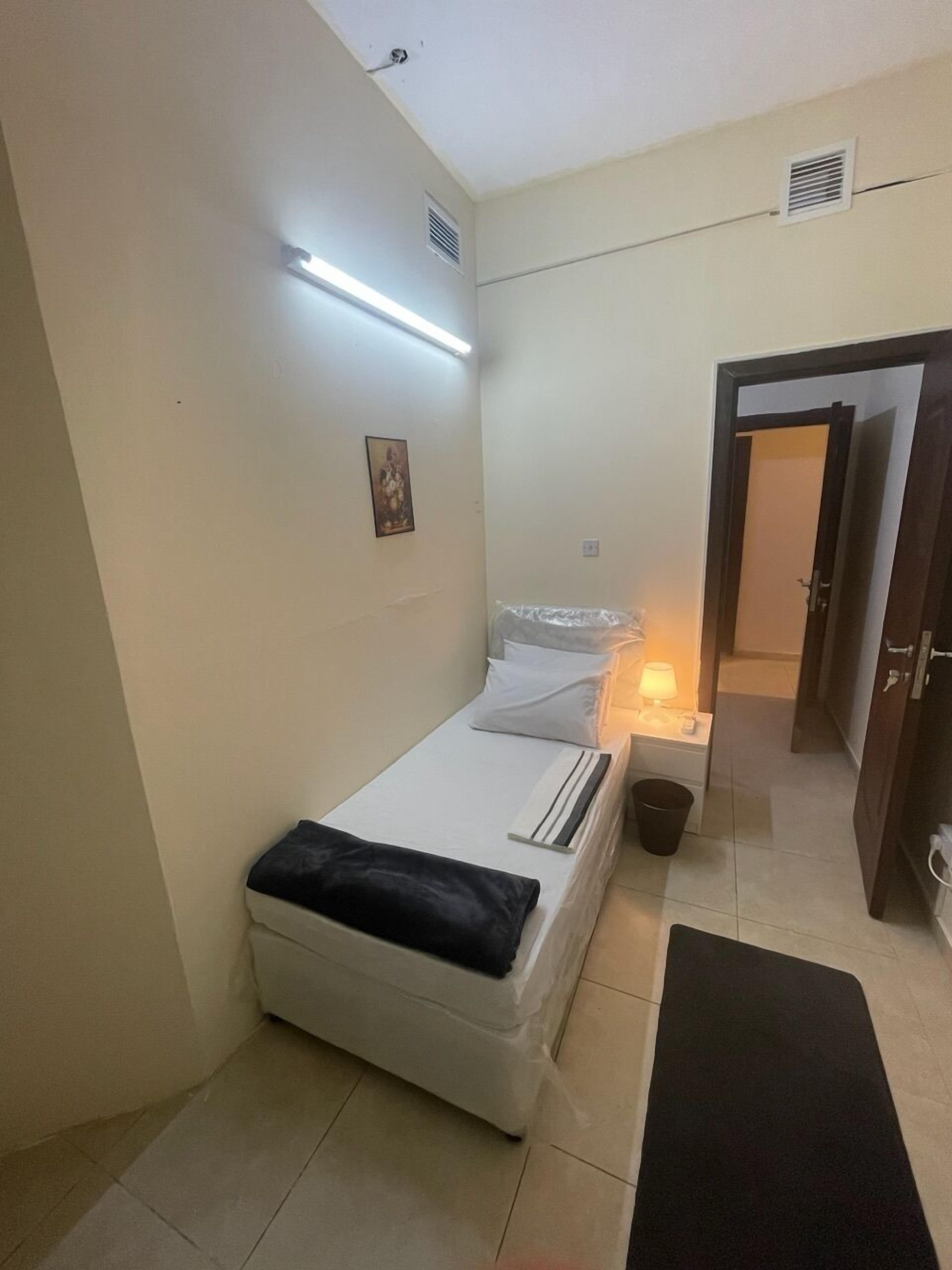 A small, neatly arranged bedroom with a single bed, a bedside table with a lamp, a small trash bin, and a door leading to another room.