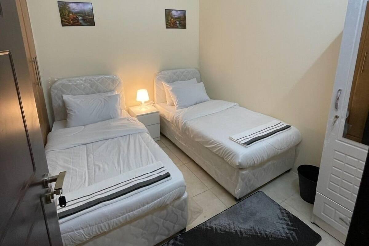 A small, clean room with two single beds, a nightstand with a lamp, a black rug, and a door with a handle.