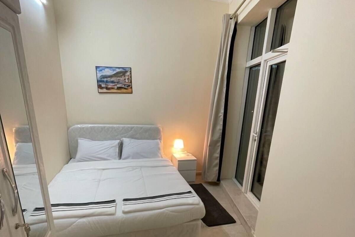 A small, neatly arranged bedroom with a bed, bedside table, lamp, and a window with curtains.