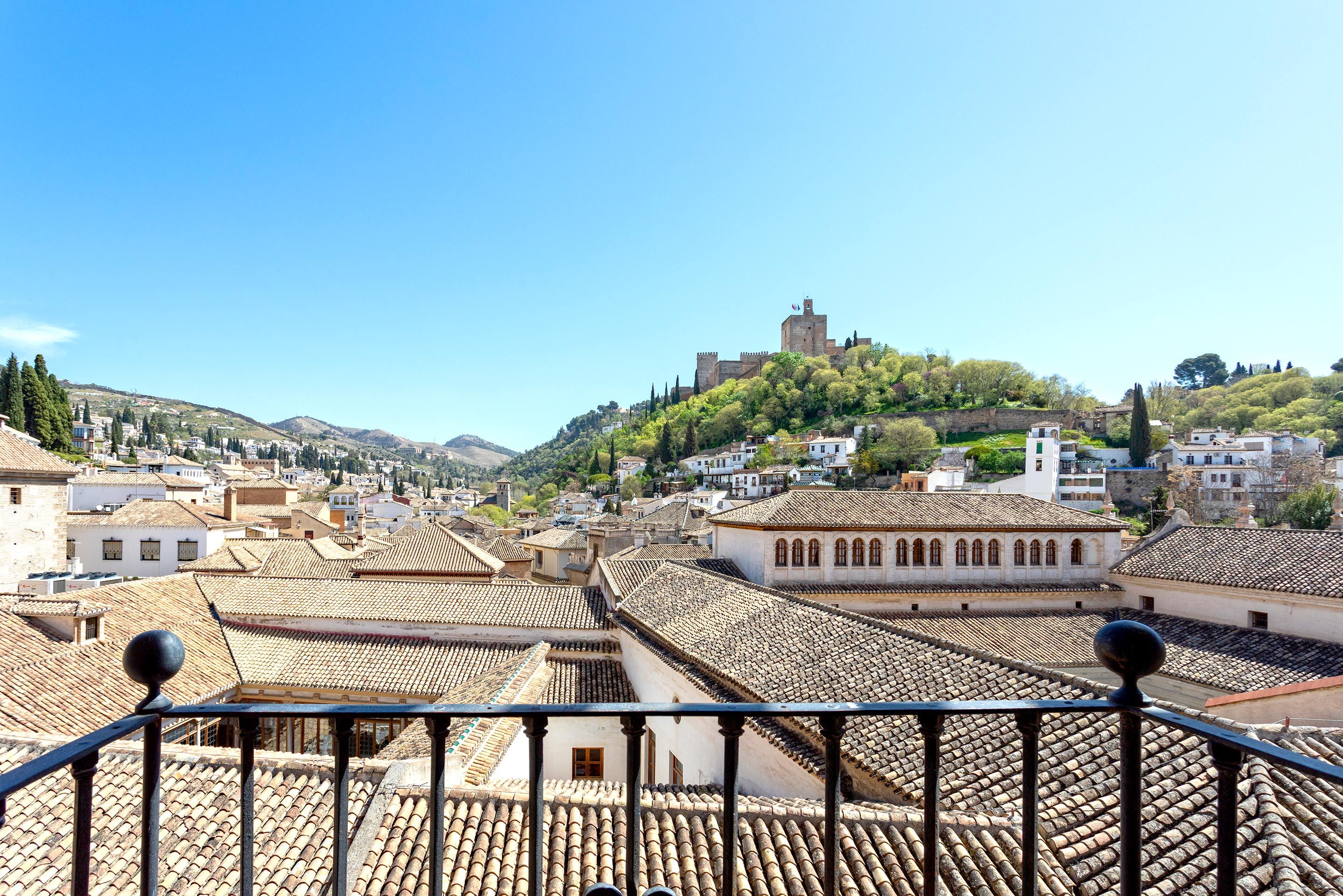 A view of a town with terracotta-tiled rooftops, a prominent castle on a hill, and a clear blue sky.
