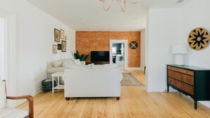 A modern living room with a brick accent wall, a white sofa, a flat-screen TV, and a wooden floor. - Newly Renovated, Vintage Inspired, Large 4 BR Home (Mount Joy)