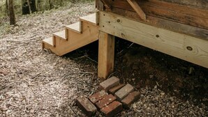 Een houten hutje met een houten trap die naar een platform leidt. Op het platform staat een witte emmer met de tekst 'w/w' en een metalen bak.