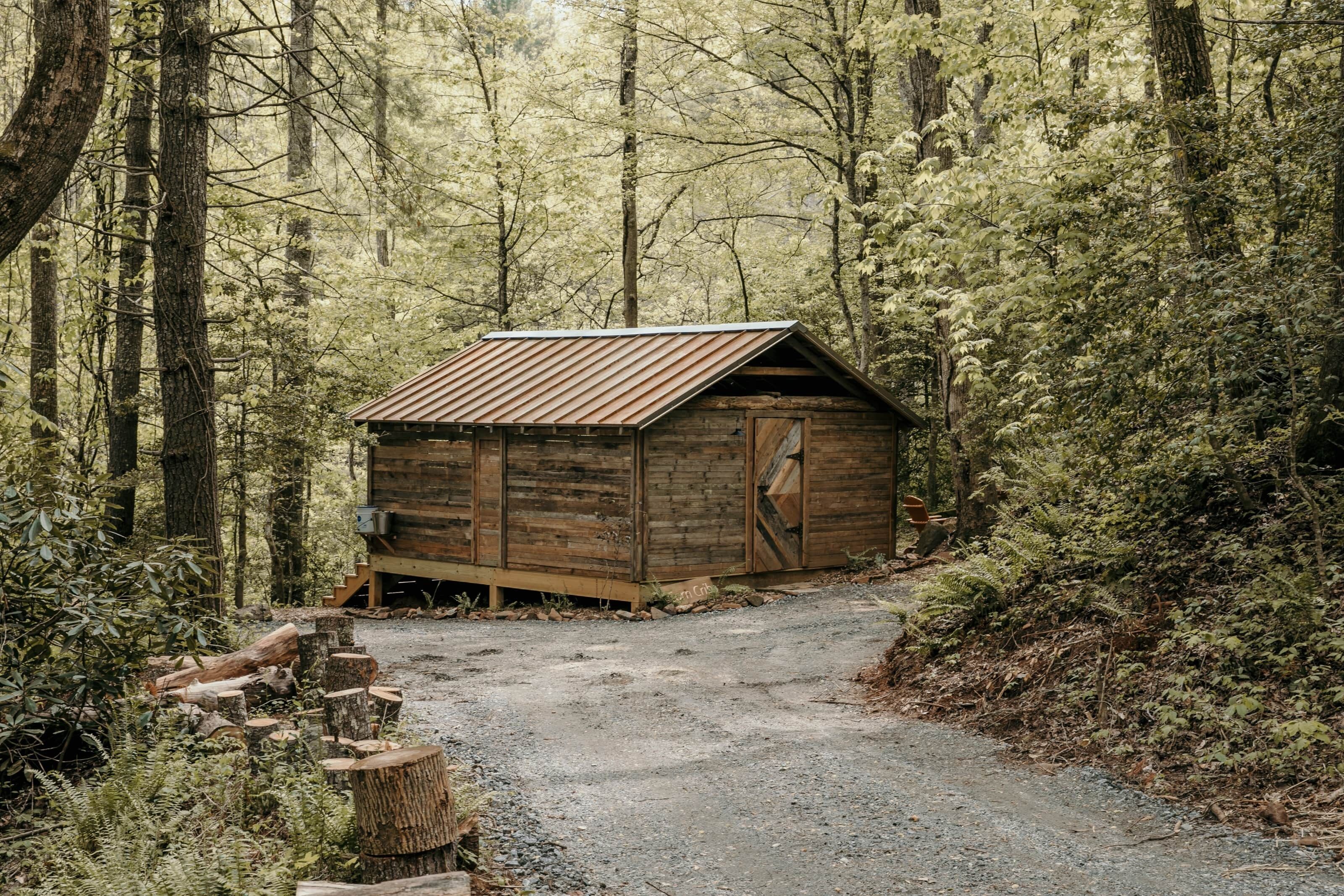 A rustic wooden cabin in a forested area with a gravel path leading to it.