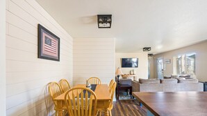 A dining area with a wooden table and chairs, a television in the background, and a sofa in the living room.