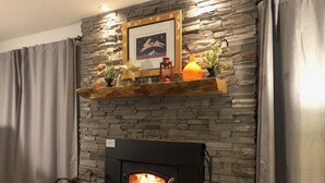 A stone fireplace with a modern design, a wooden shelf above it holding decorative items, and a framed picture on the wall. - Walk to Sisters | Hot Tub, Pool + Fireplace (Sisters)
