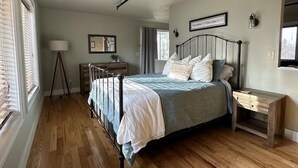 Een slaapkamer met een metalen bed, nachtkastje, lamp en een raam met jaloezieën.