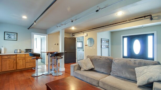 Una sala de estar moderna con sofá, mesa de centro y taburetes de bar. Hay una zona de cocina con armarios de madera y una mesa de comedor.