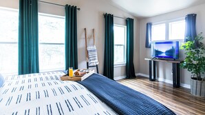 Habitación de hotel con cama, televisor sobre un pedestal, una planta y ventana con cortinas.