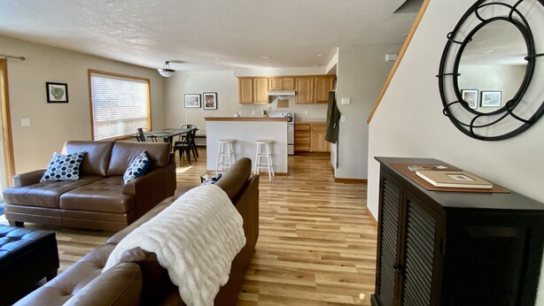 A modern living room with a brown leather sofa, a white throw blanket, and a fireplace. A kitchen with wooden cabinets and a bar area is visible in the background.