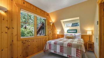 Een slaapkamer met een groot raam, houten wanden, een gebogen beddegoed, twee nachtkastjes met lampen en een ingelijst kunstwerk aan de muur.