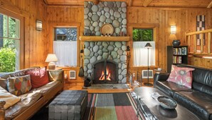 A cozy cabin interior with a stone fireplace, wooden walls, leather sofas, and a bookshelf.