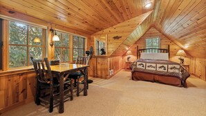 Une cabane en rondins comprenant un coin repas, une chambre avec un grand lit et de nombreuses fenêtres donnant sur la forêt.