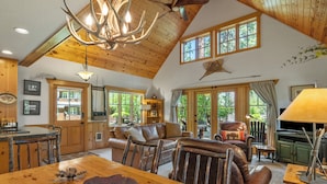 A spacious living room with a high ceiling, wooden beams, a large dining table, leather furniture, and large windows offering a view of the outdoors.