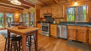 A wooden cabin-style kitchen with a wooden table and bar stools, a stove, oven, dishwasher, and large windows with curtains.