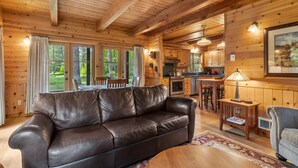 A wooden cabin interior with a leather sofa, dining table, kitchen area, and large windows.