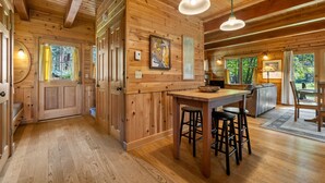 A wooden cabin interior with a kitchen area, dining table, bar stools, and large windows.